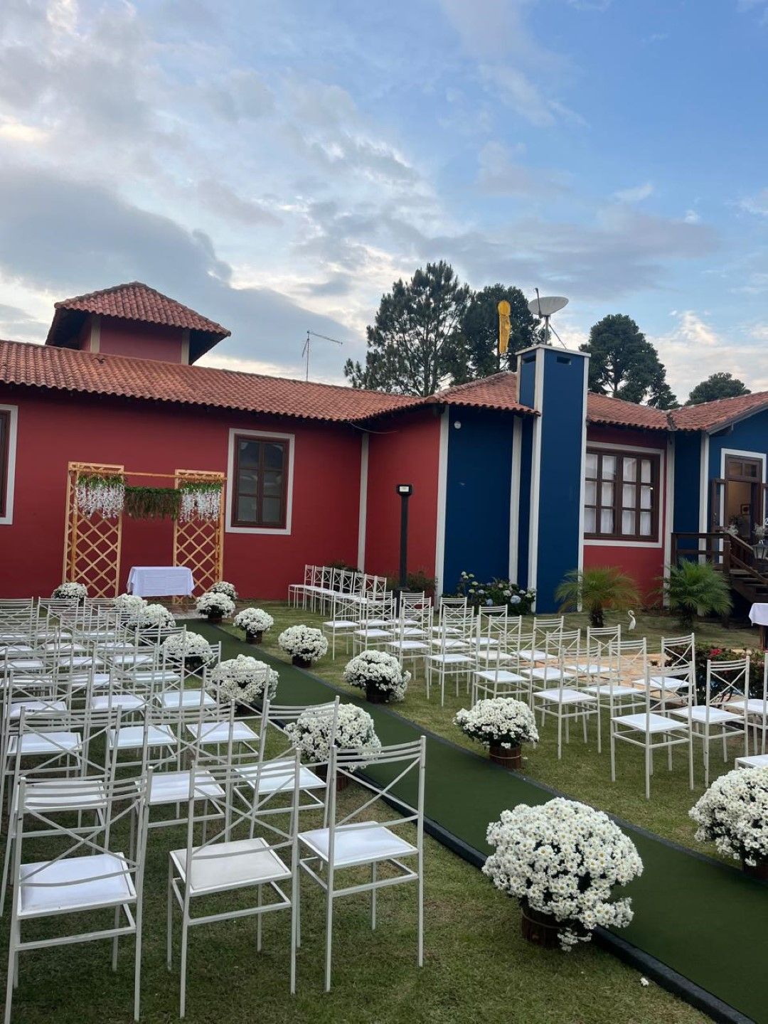 Cerimônia de casamento ao ar livre com cadeiras brancas, flores e prédio vermelho/azul.