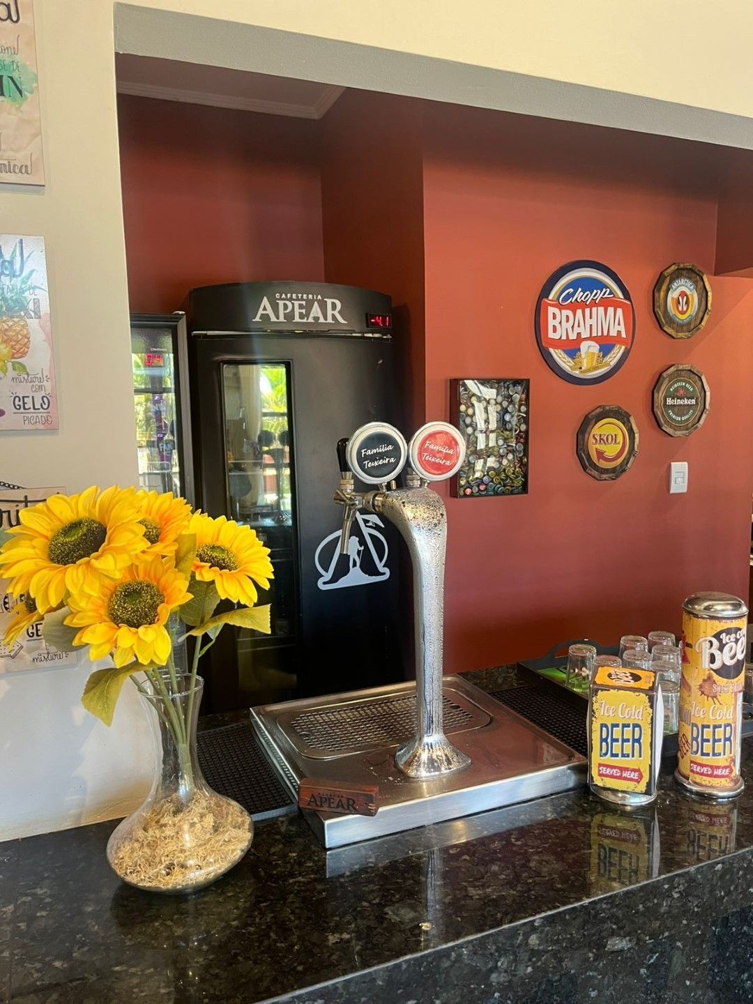 Um bar com torneira de cerveja, girassóis e latas de cerveja no balcão, com um cooler e rótulos de cerveja na parede.