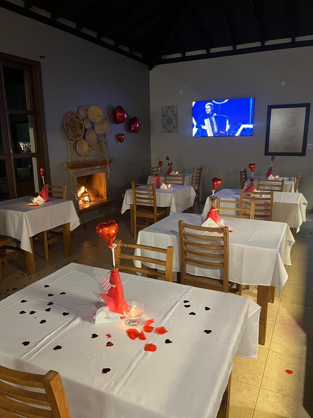 Mesas de restaurante preparadas para um jantar romântico, lareira, TV, centro de mesa com rosas vermelhas e pétalas de rosas espalhadas.