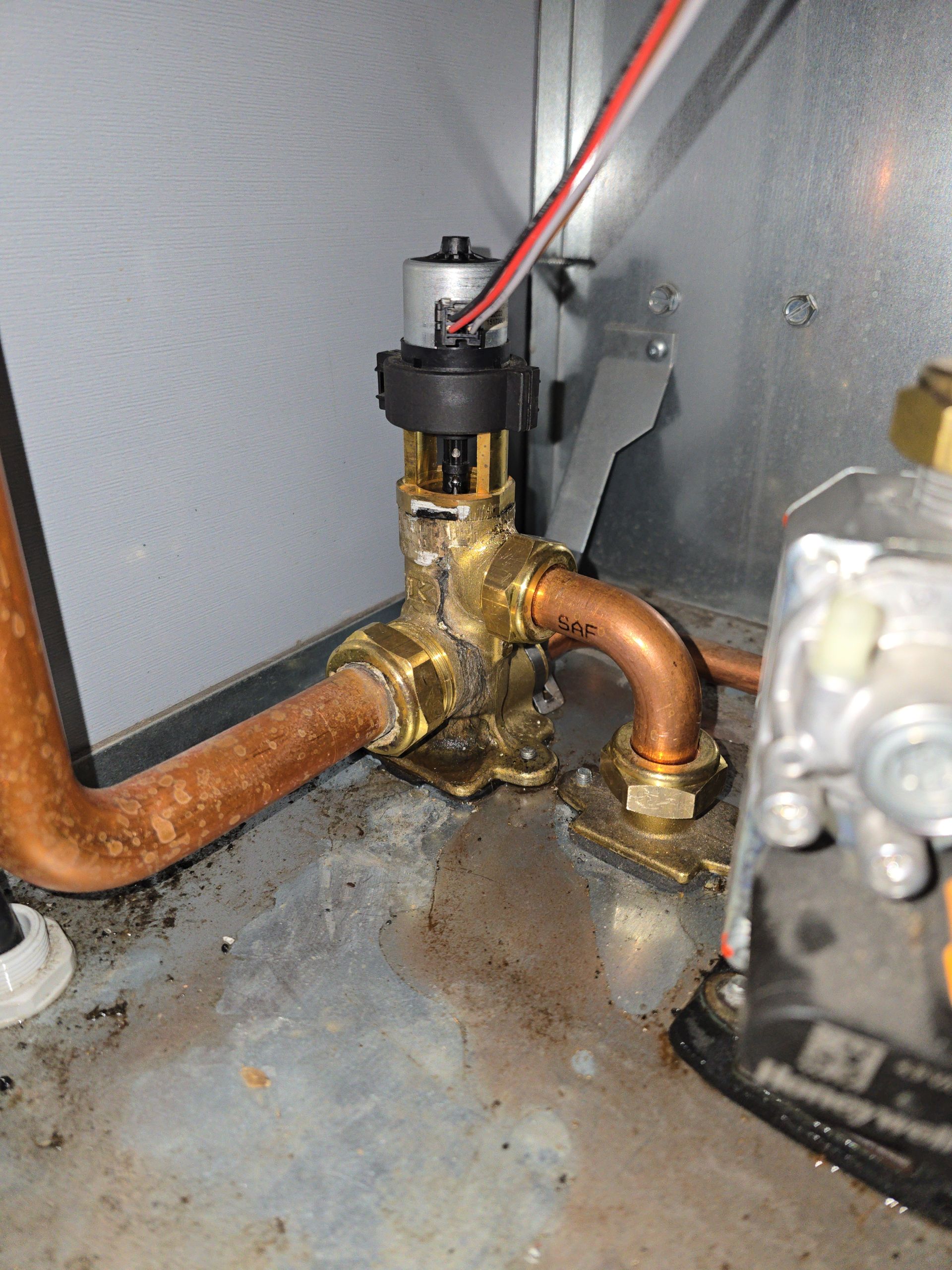 A brass valve with copper pipes, likely part of a furnace, inside a metal cabinet. Wires are connected.