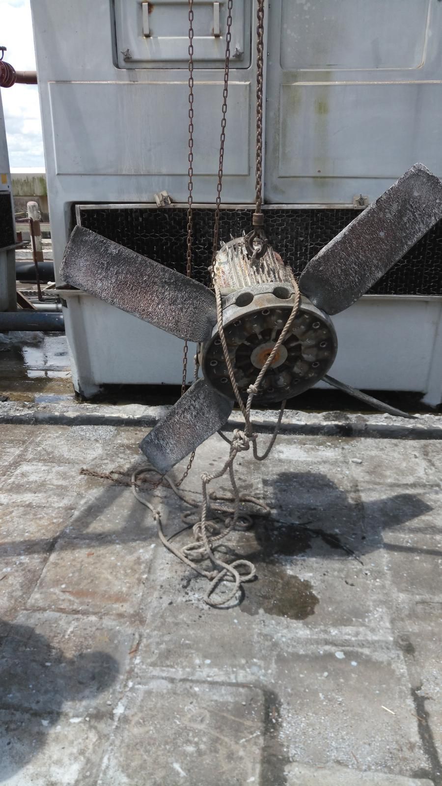 Damaged HVAC fan hanging by chains on a rooftop, blades covered in debris, against a large unit.