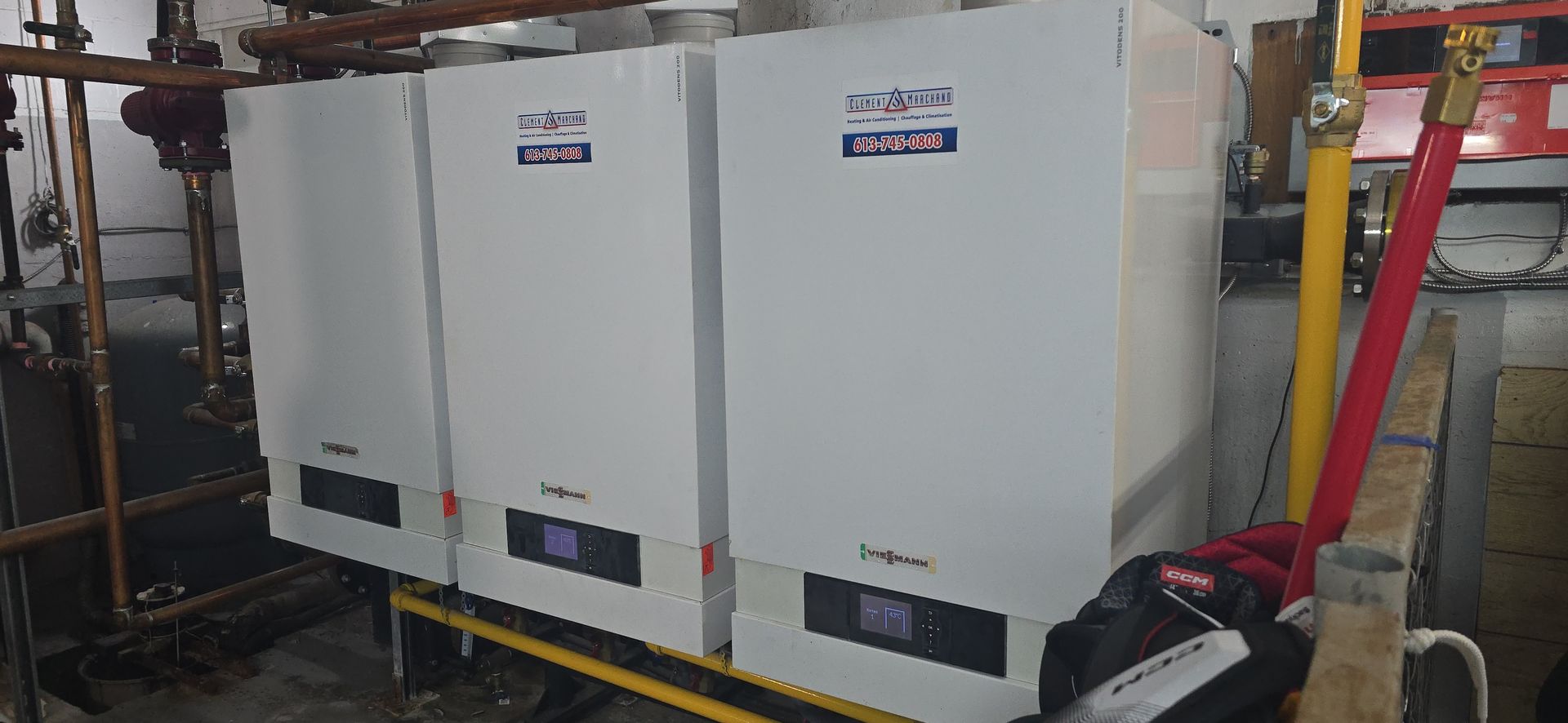 Three white water heaters are in a utility room. Copper piping is overhead. A yellow and red pipe stand in front.