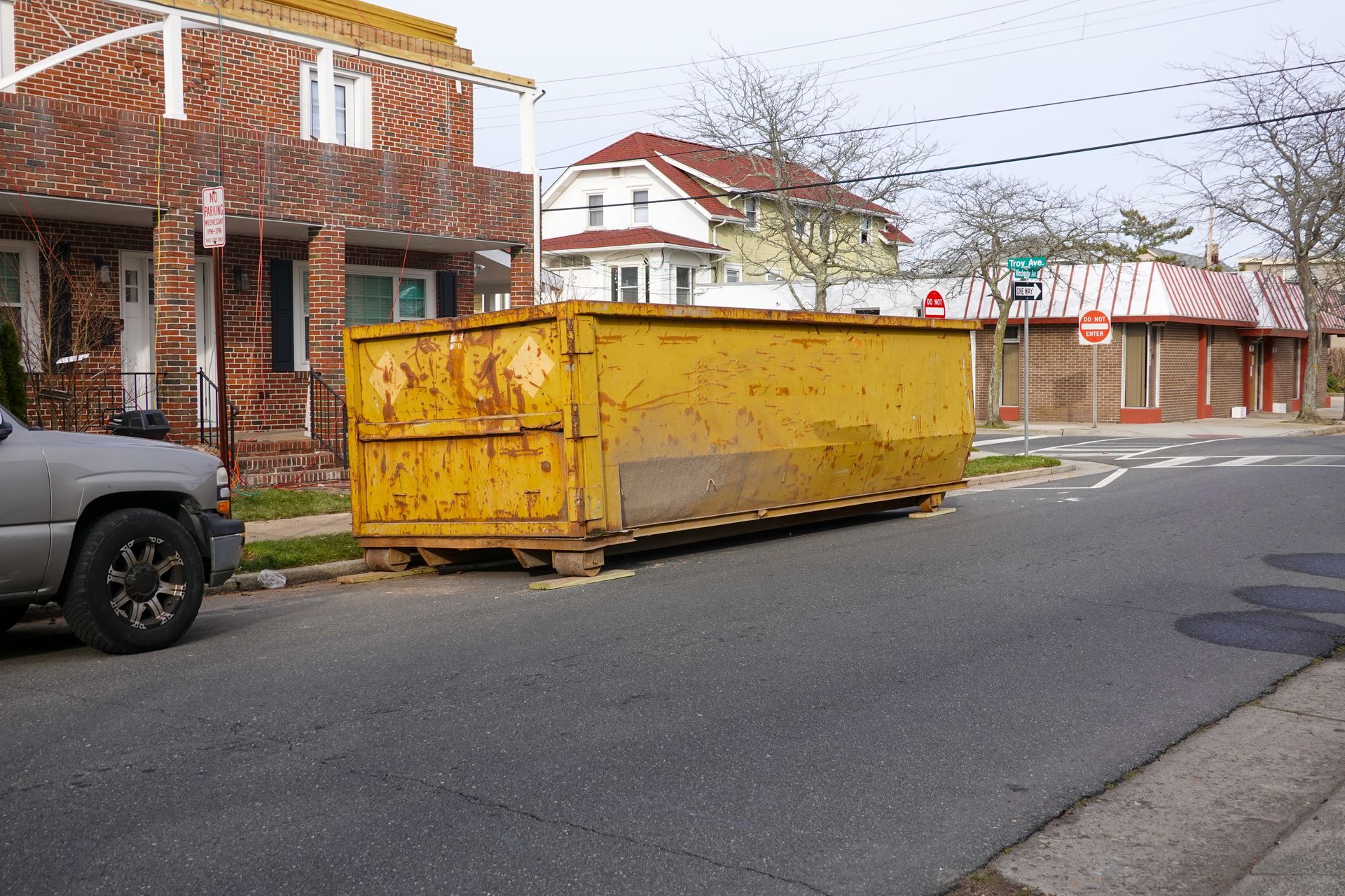 Yellow dumpster on the side of a street in front of a brick building and a residential house. Yellow dumpster on the side of a street in front of a brick building and a residential house.