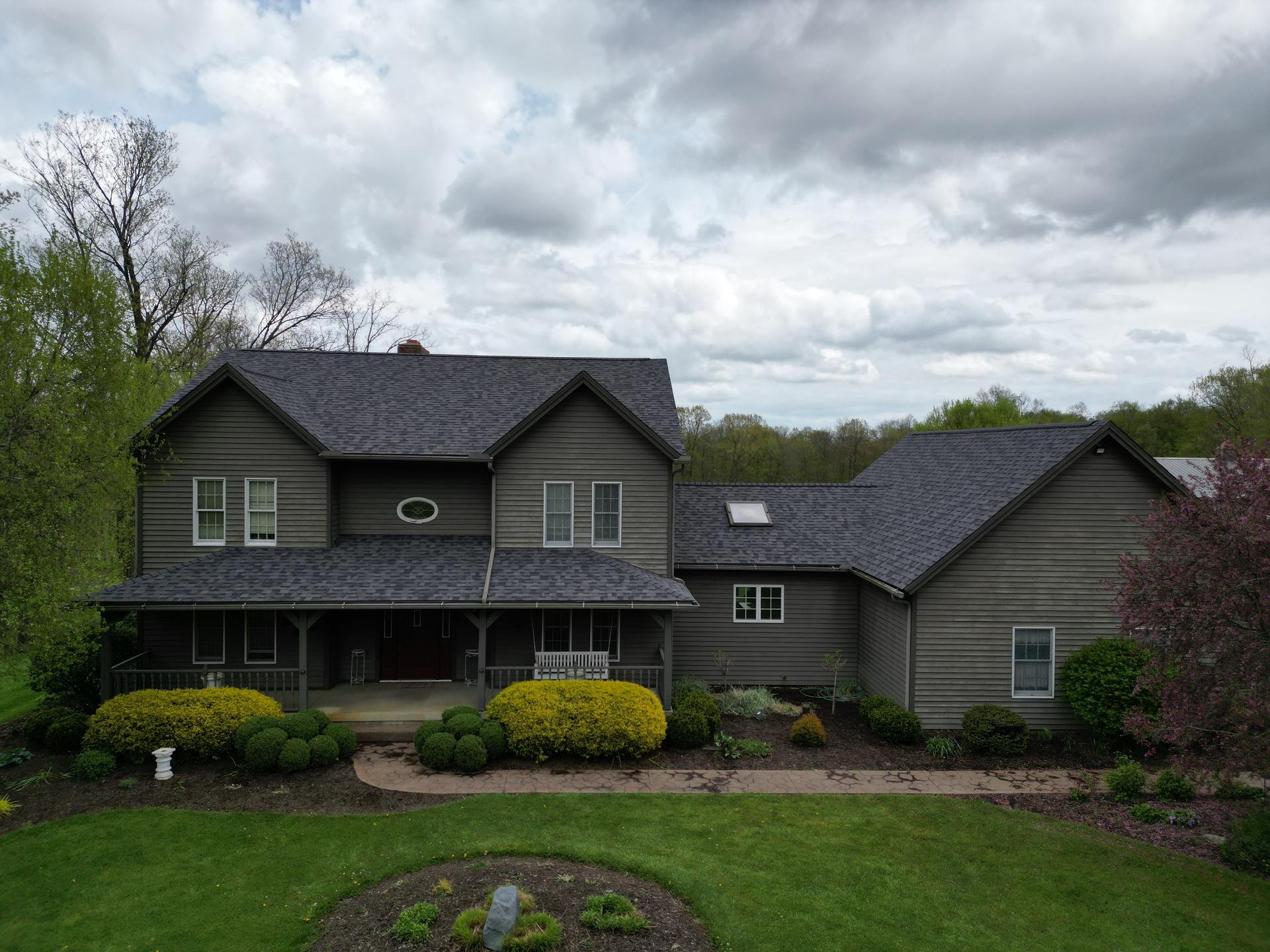 Two-story gray house with a dark roof, porch, and landscaping under a cloudy sky.