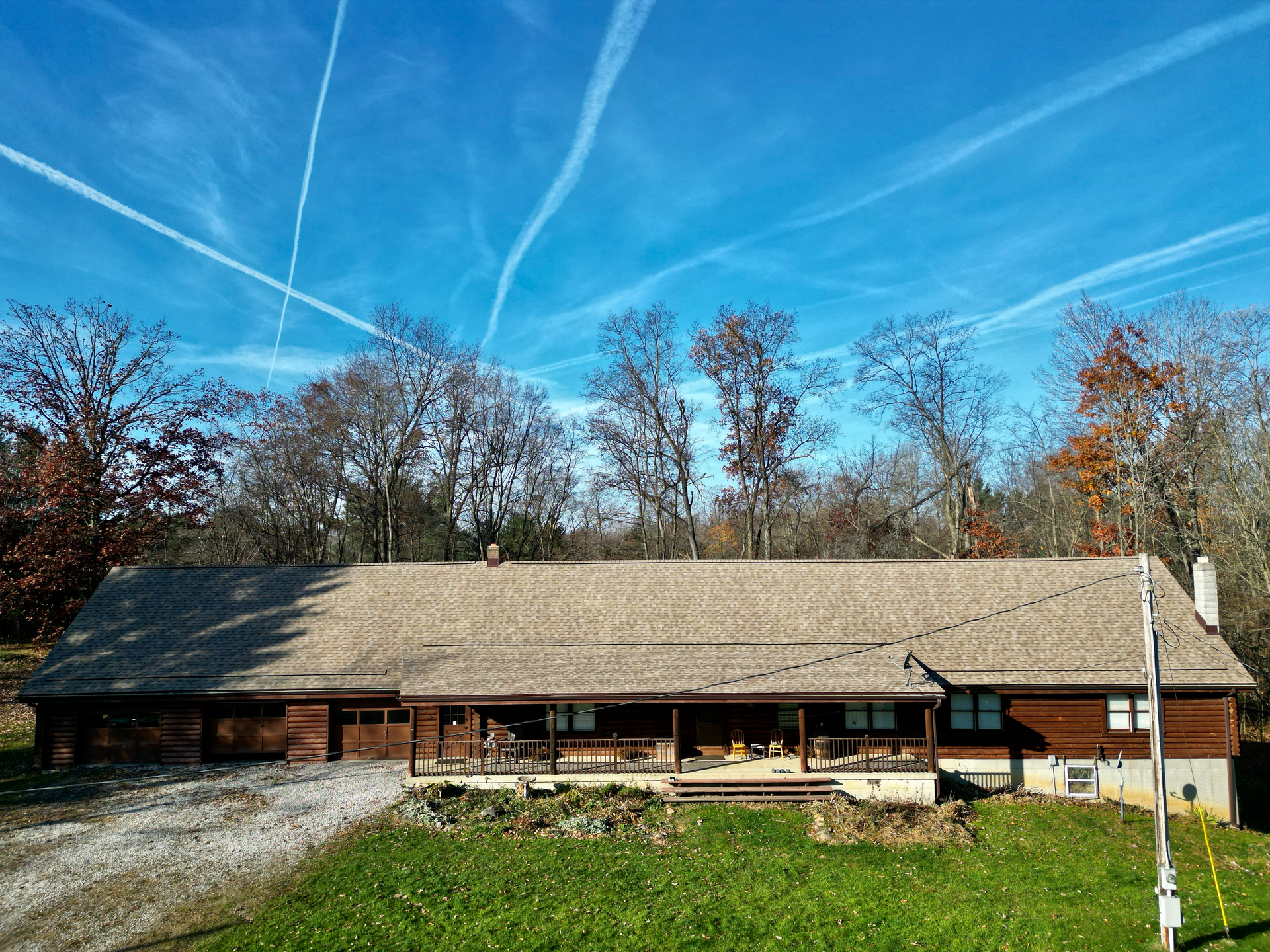 A long, single-story cabin with a wide porch in a grassy area, surrounded by trees under a blue sky with contrails.