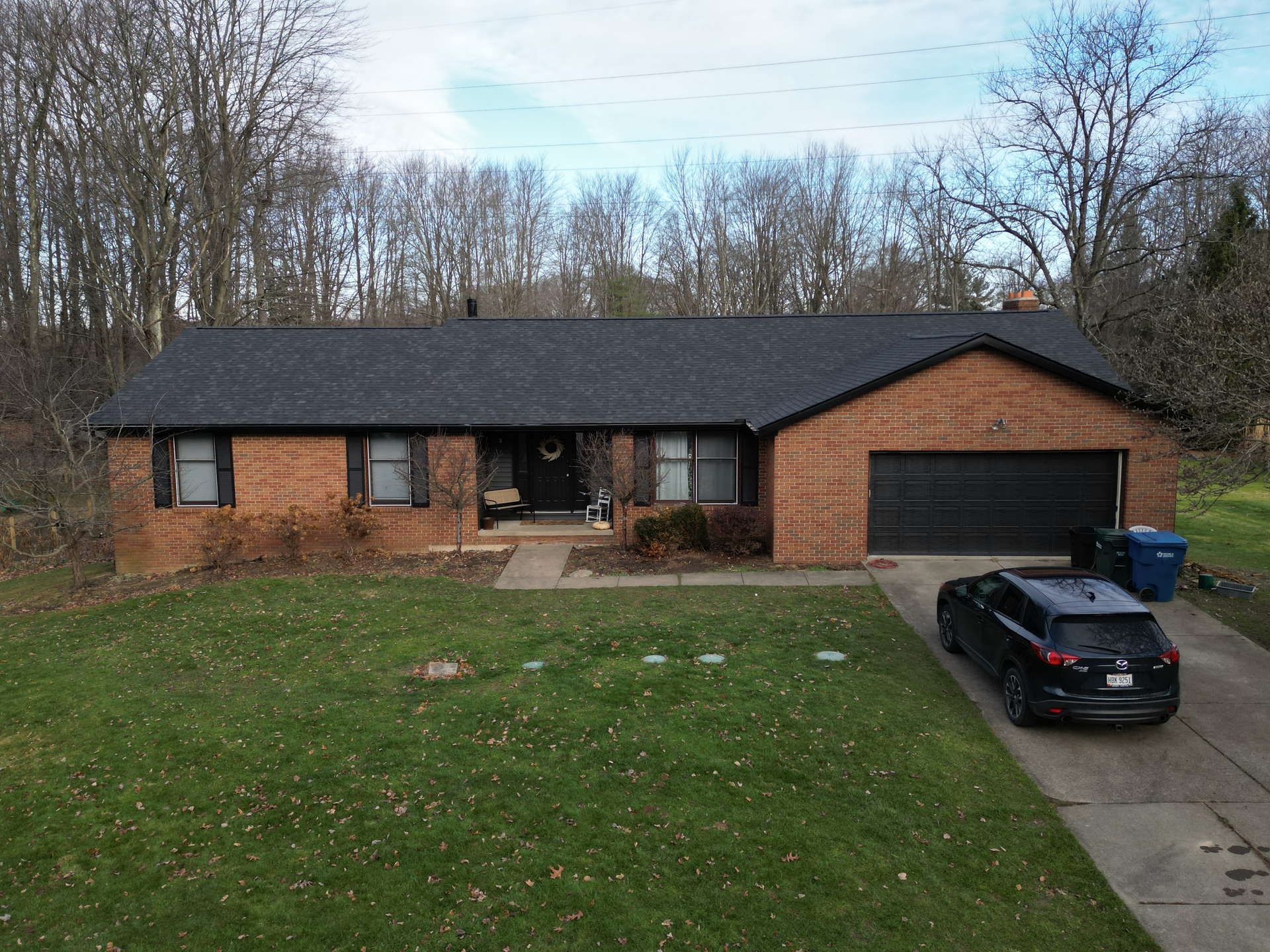 Brick ranch house with black roof and garage door, car parked in driveway, blue trash bin. Green grass lawn.