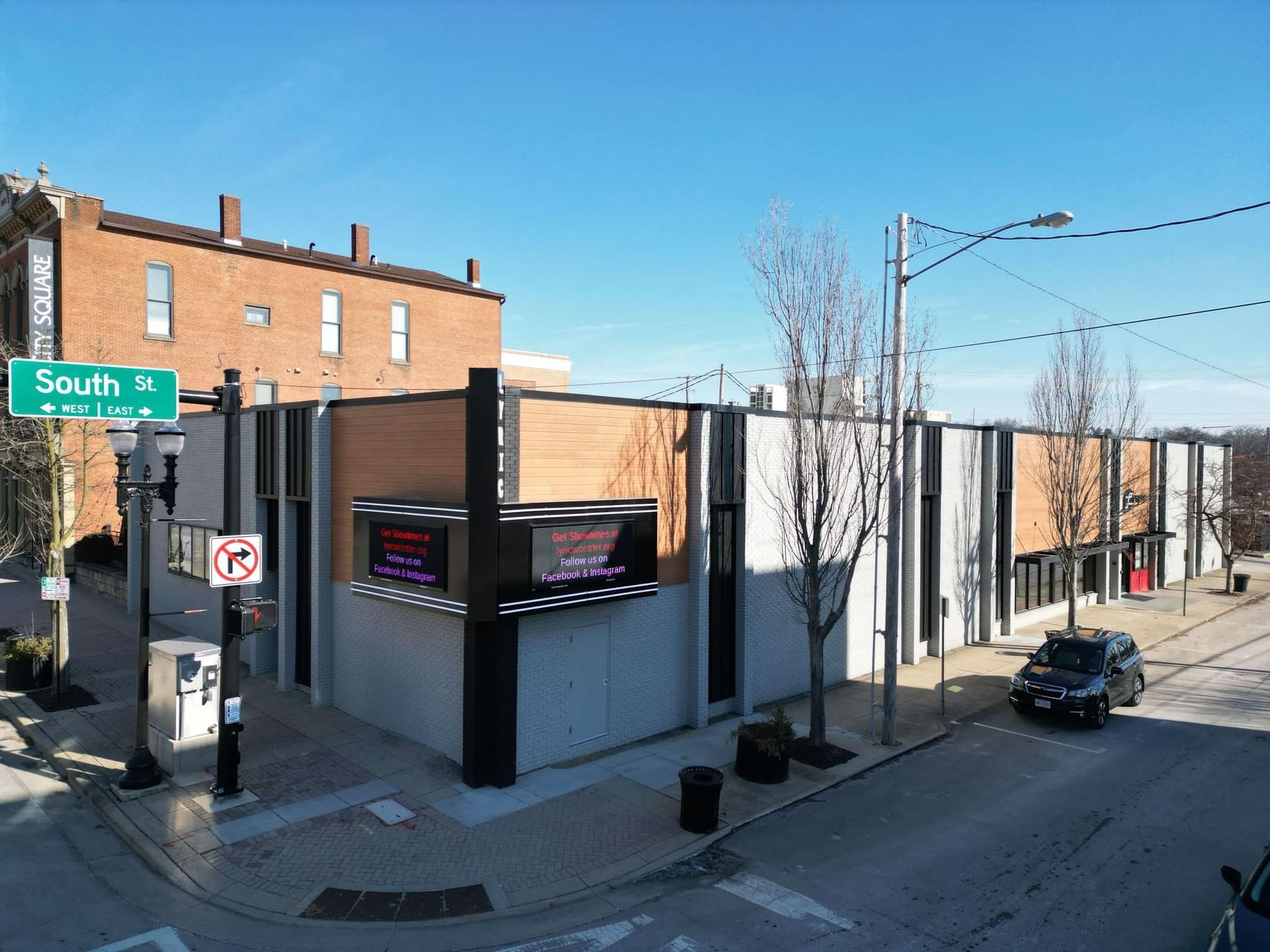 Exterior view of a modern building on a street corner with a large digital display. A black vehicle drives on the street.