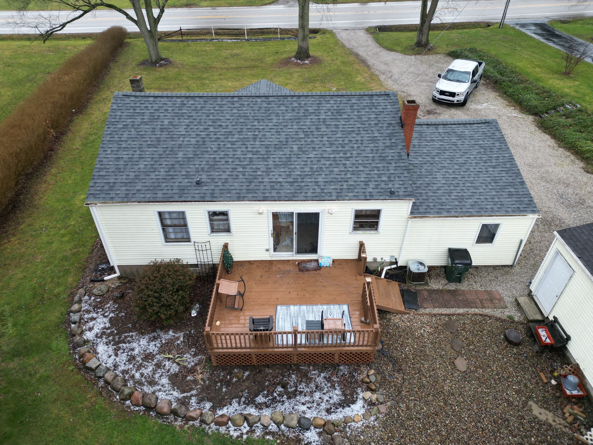 Overhead view of a one-story house with a wooden deck in front. A white truck is parked on the gravel driveway.