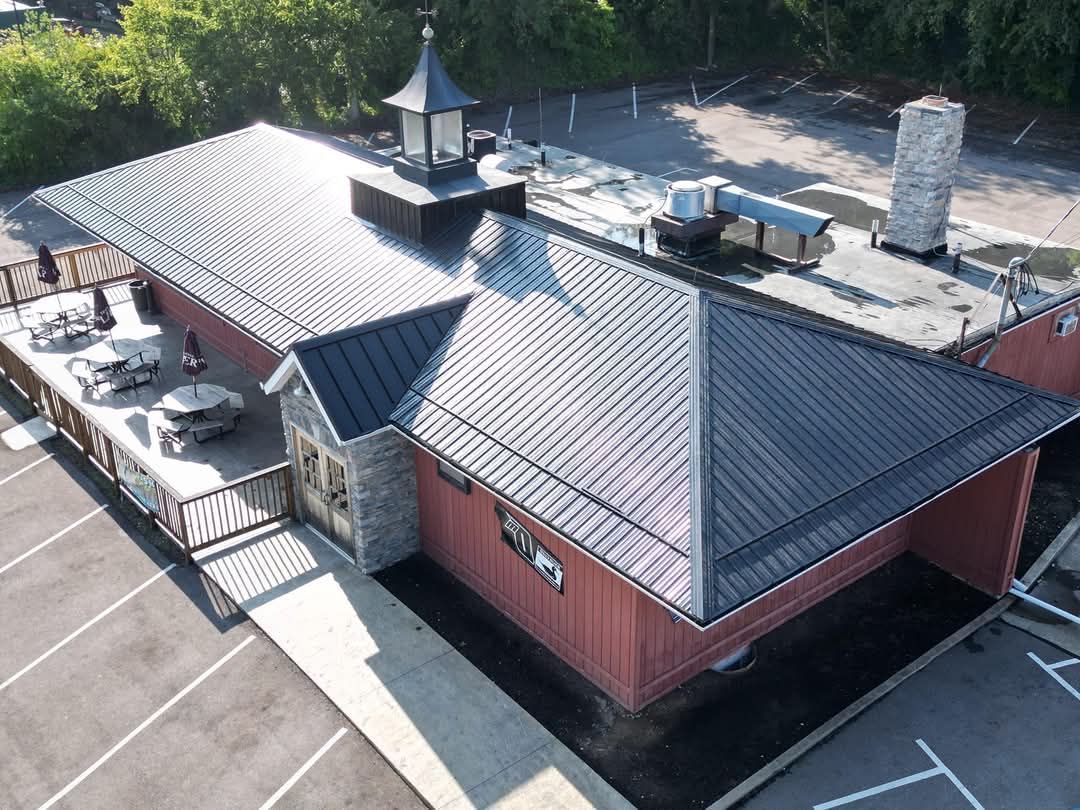 Aerial view of a red building with a black metal roof, a small deck with tables, and a surrounding parking lot.