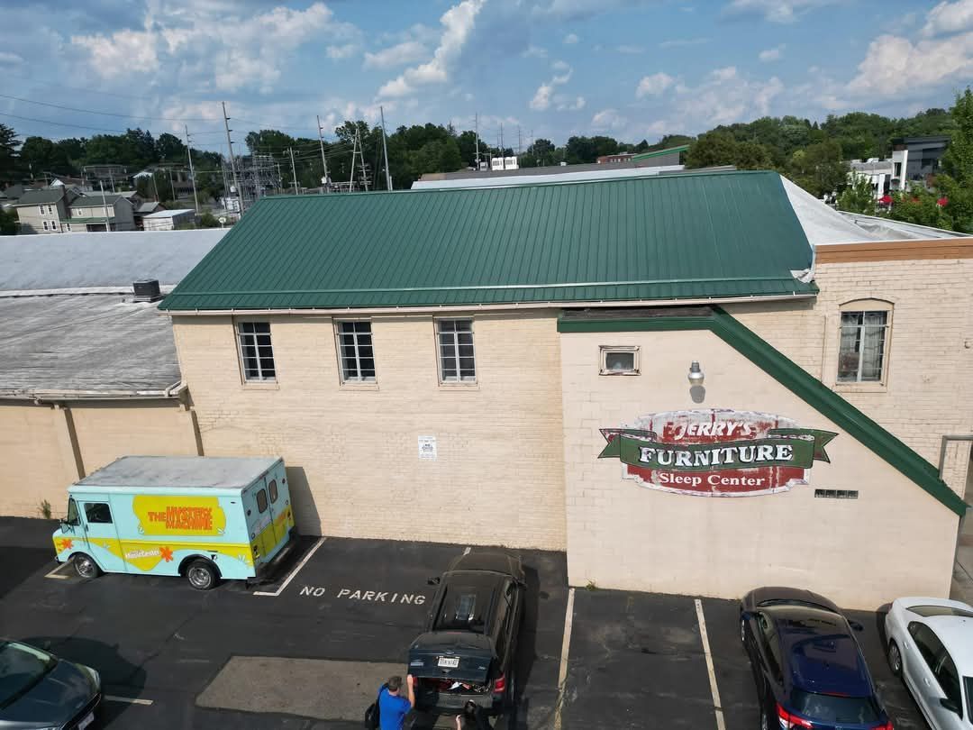 Exterior view of a furniture store with a green roof and a food truck parked beside it. Parking lot with several cars.