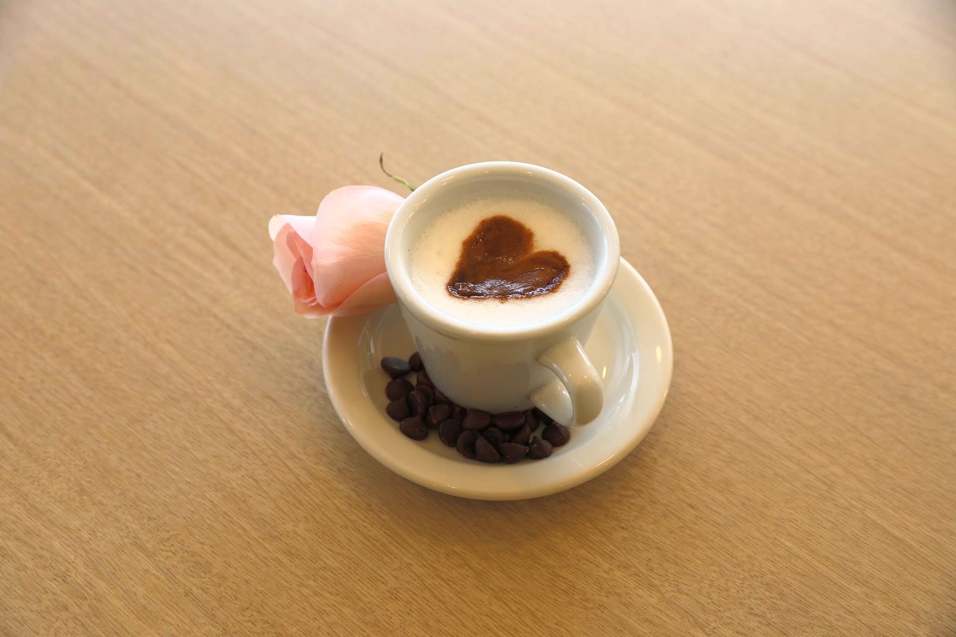 A latte with a heart drawn on it and coffee beans on a saucer with a pink rose. 