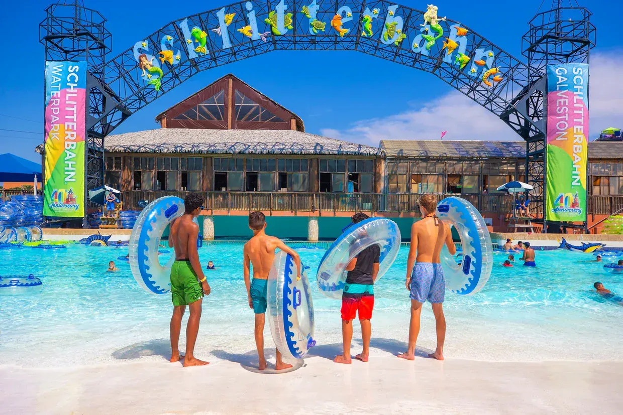 A group of teenage boys are standing in front of a wave pool holding rafts.