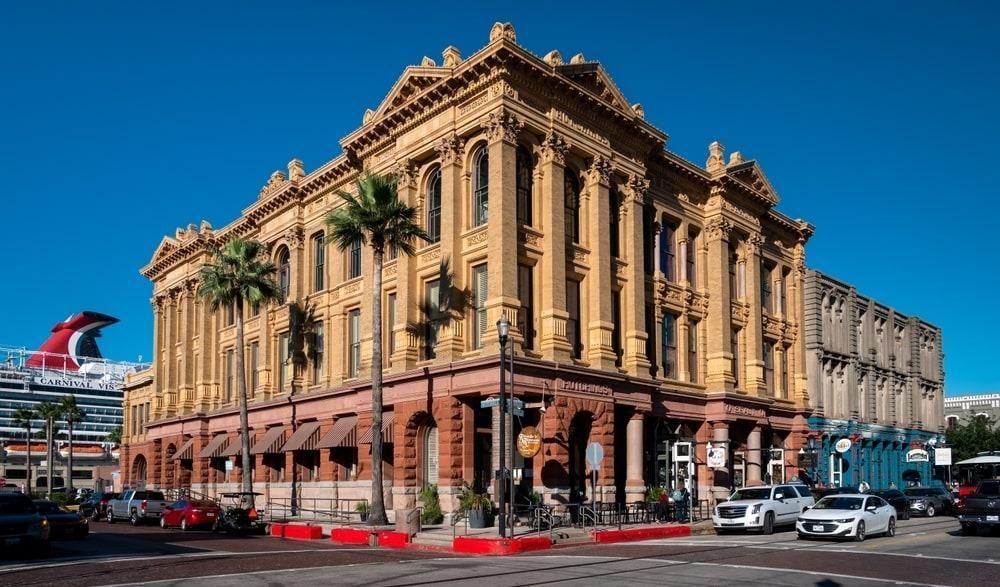 A large building with cars parked in front of it and a cruise ship in the background.