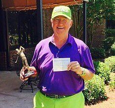 A man in a purple shirt and green pants is holding a trophy and a check.