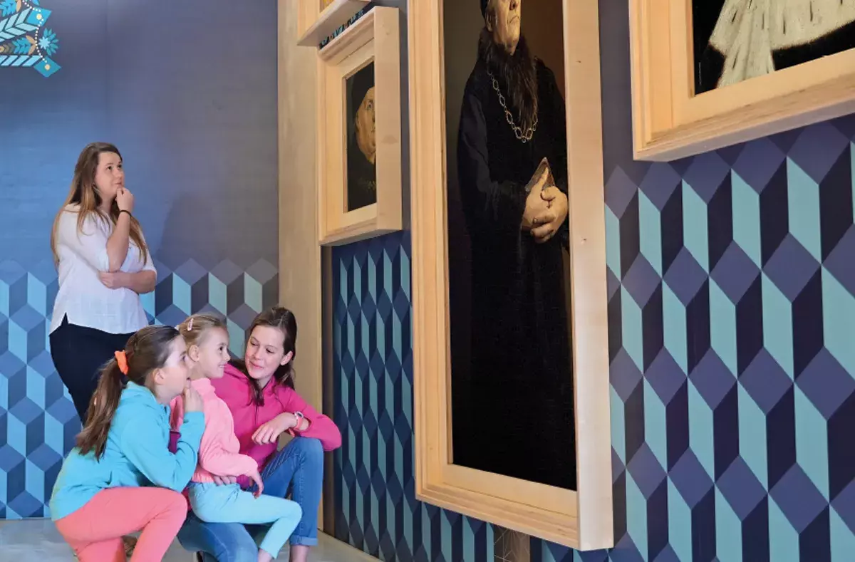 Un adulte et trois enfants admirent une peinture dans une salle aux murs ornés d’un motif géométrique bleu.