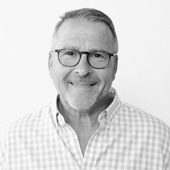 A man wearing glasses and a plaid shirt is smiling in a black and white photo.