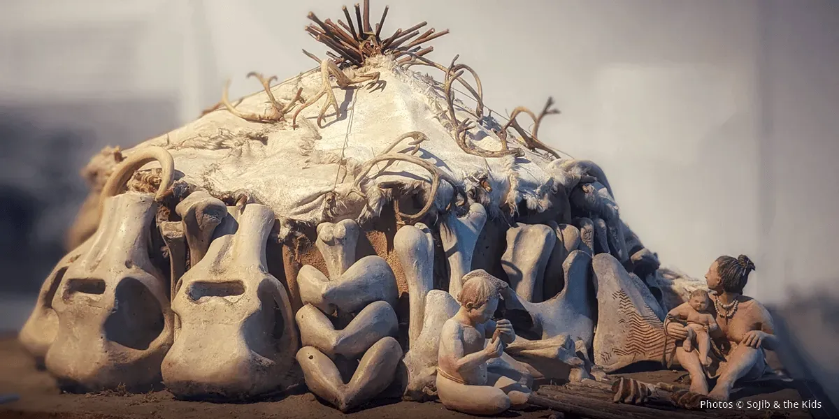 Maquette de hutte avec des personnages assis à l’avant et entourée de gros os.