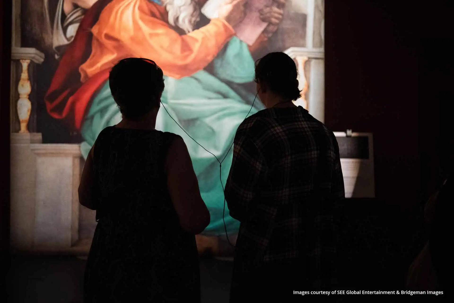 Deux personnes partagent des écouteurs connectés à un Podcatcher devant une peinture de l’exposition sur la chapelle Sixtine.