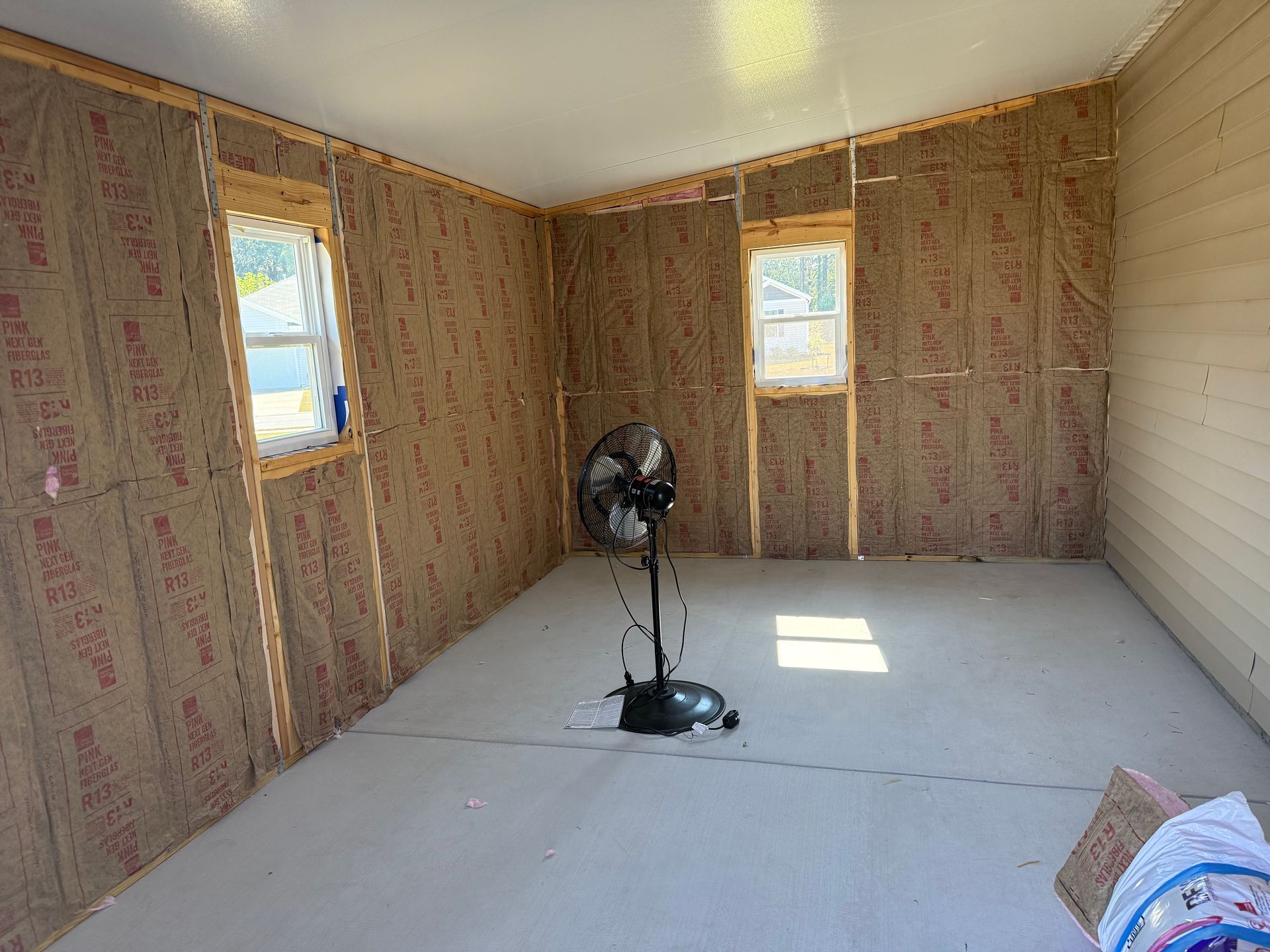 Interior of a room under construction with exposed insulation, two windows, and a fan.