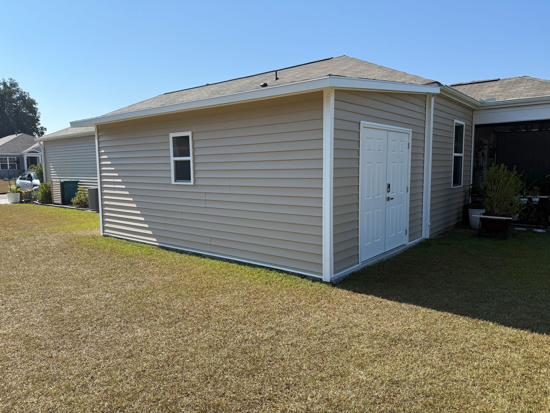 Tan shed with white trim, a small window, and double doors on a grassy lawn under a clear blue sky.