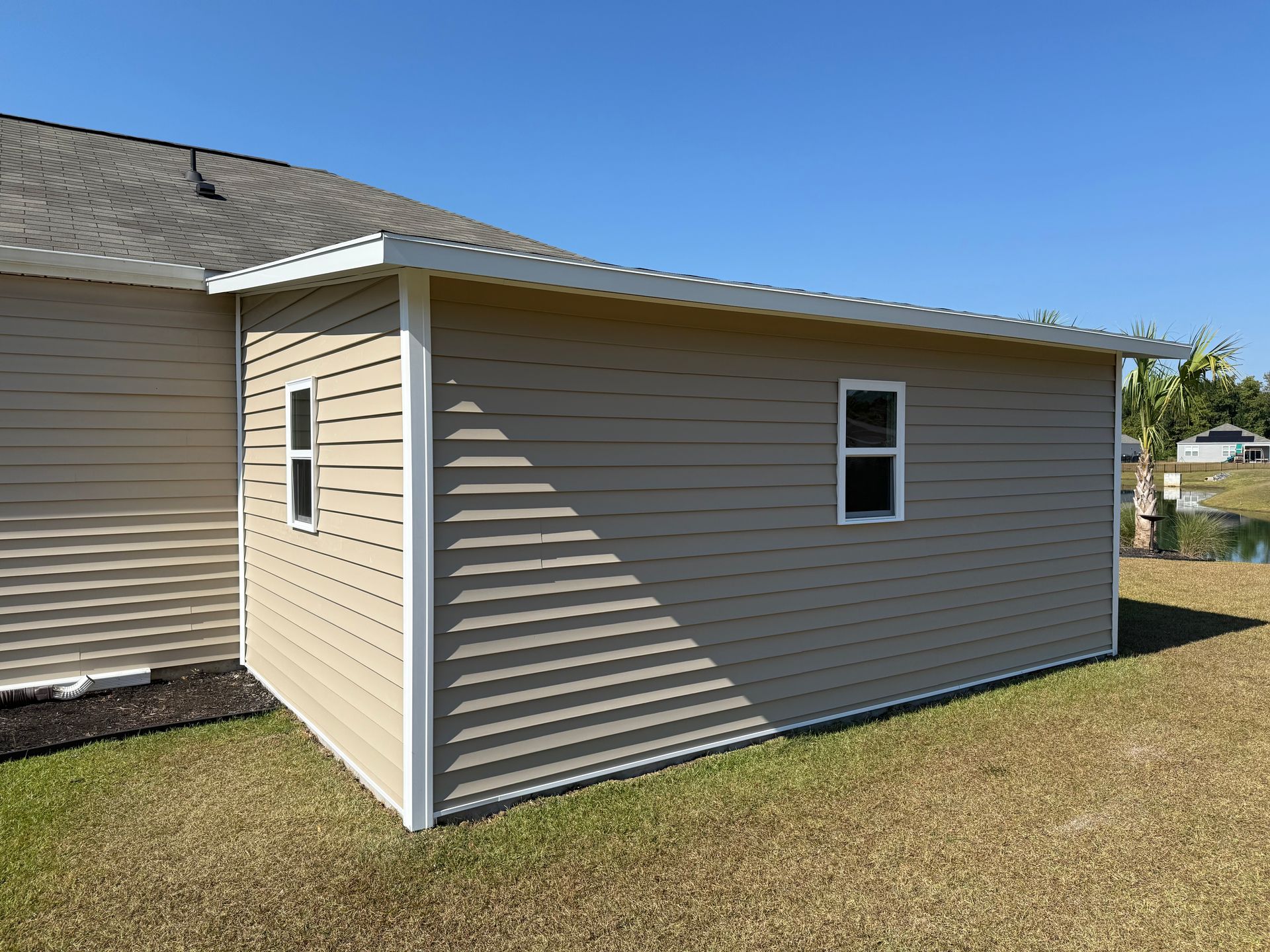 Tan building with white trim, two windows, and a metal roof against a blue sky.