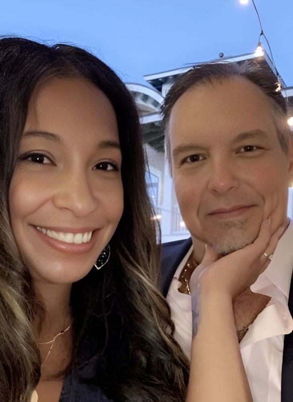 Woman smiling, resting hand on face, beside a man with graying hair, both close together outdoors.