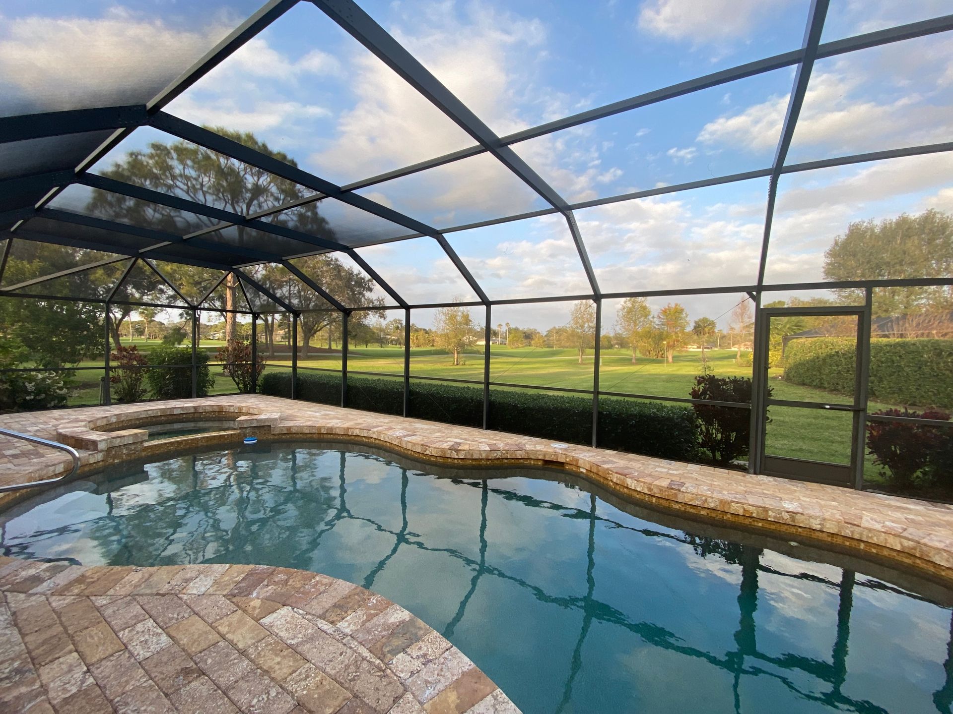 Pool and hot tub under screened enclosure, overlooking a green lawn under a partly cloudy sky.