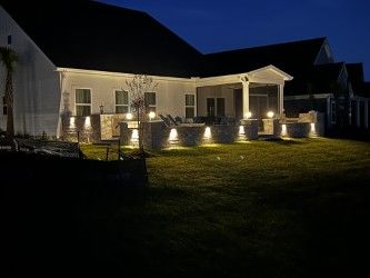 Backyard at night, lit by landscape lighting. House with a covered porch, stone features, and a lawn.