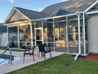 Screened-in porch beside a pool. White frame with chairs and table. Green lawn, blue sky.