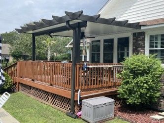 Wooden deck with dark pergola. House in background. Green grass and shrubs.