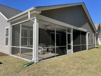 Screened-in porch attached to a house with gray siding. White trim, seating, and a ceiling fan are visible inside.