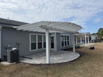 Backyard patio with pergola attached to a gray house, featuring a brick patio and a cloudy sky.