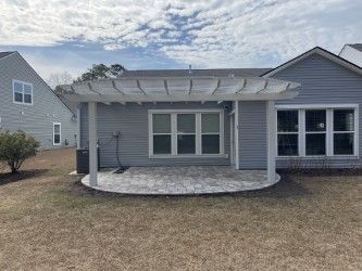 Backyard patio with pergola, gray siding, and a stone patio. Overcast sky.