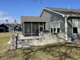 Screened-in porch attached to a house with gray siding. Low stone wall, clear sky.
