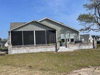 A light blue house with a screened porch and small brick patio on a grassy lawn under a clear sky.