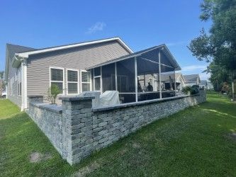 Screened porch attached to a light gray house, with a low brick retaining wall. Blue sky.