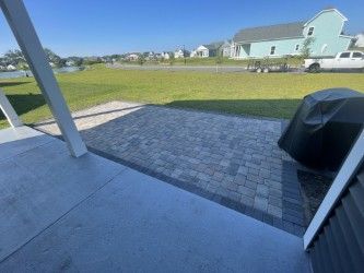 Patio with pavers and a covered grill, overlooking a grassy area and houses under a clear blue sky.