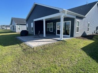 Backyard patio with a covered seating area. Dark blue house, grey pavers, and green lawn on a sunny day.