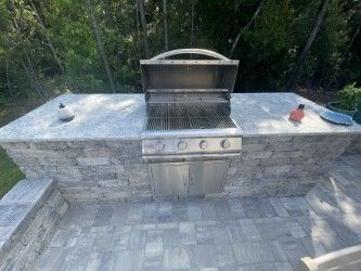 Built-in outdoor kitchen with a stainless steel grill, gray stone counters, and matching base in a backyard setting.