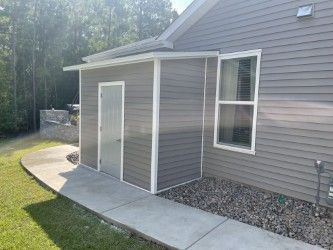 Gray shed attached to a gray house, with a concrete walkway and window.