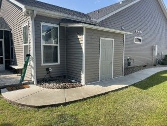 Exterior view of a gray shed attached to a house with a concrete walkway in a yard.