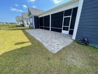 Screened-in patio with gray brick pavers, surrounded by grass, adjacent to a blue house on a sunny day.
