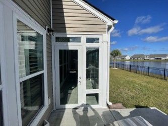 Exterior view: white-framed glass doors and window on a tan siding house, overlooking a yard and lake under a blue sky.