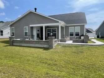 Gray house with a covered patio, low brick wall, and green lawn on a sunny day.
