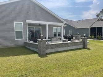 Backyard patio with a retaining wall and covered seating area, next to a gray house, on a sunny day.