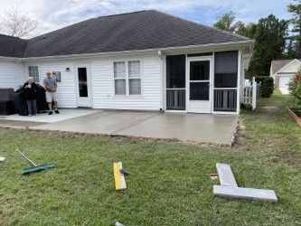 Backyard with new concrete patio; two people stand near a screen-enclosed porch; tools on the grass.