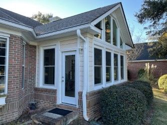 White-walled sunroom addition with a brick base. Glass windows and a door are featured. Green bushes in front.