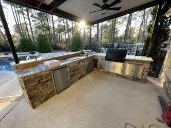 Outdoor kitchen with stone counters, grill, fridge, and sink under a covered patio.