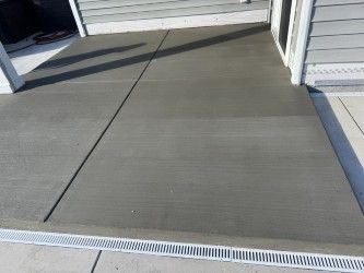 Newly poured gray concrete patio with linear drainage along the edge, next to a house.