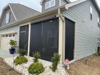 Screened porch attached to a light blue house with tan trim. Landscaping includes small bushes and white rocks.